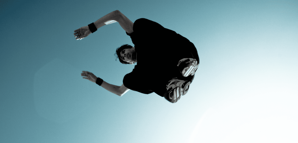 Sprung eines Sportlers mit ausgestreckten Armen vor hellem Himmel, während er sich in der Luft dreht.